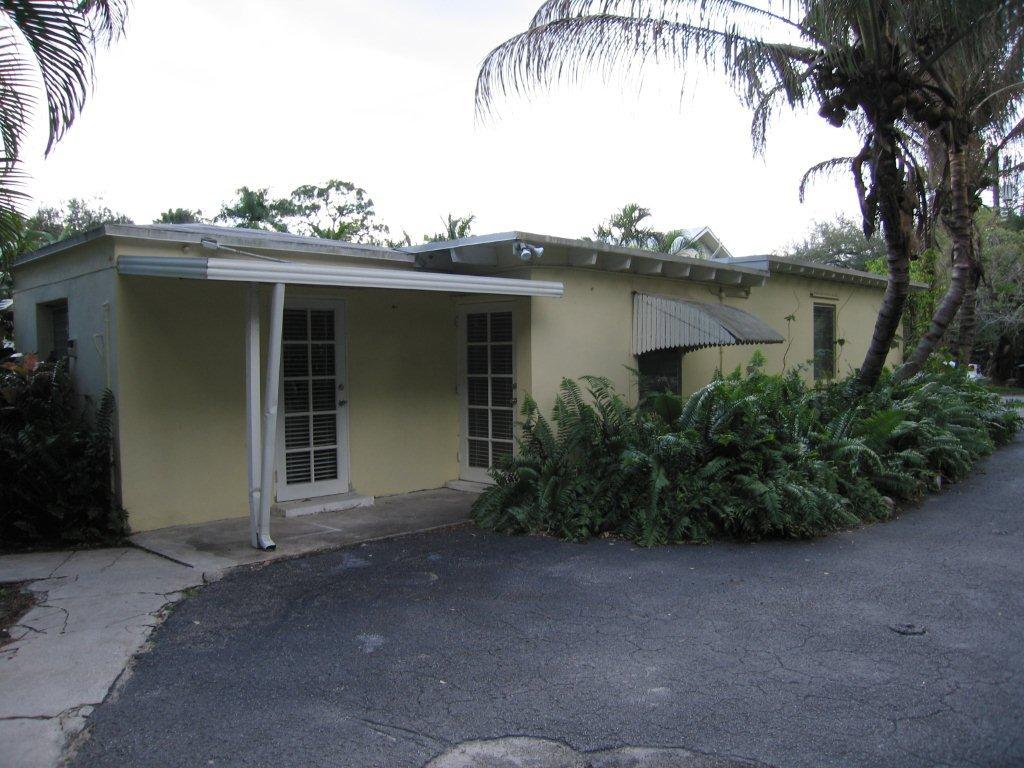 Small, single-story beige house with a driveway and greenery.