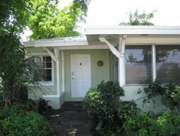 Small green house with white trim and a front door surrounded by bushes.